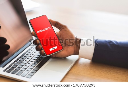 Safety Concept. Male hand holding smart phone with emergency number on red screen over office background and laptop Royalty-Free Stock Photo #1709750293