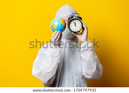 Medic in protection suit and mask hold Earth globe and alarm clock on yellow background