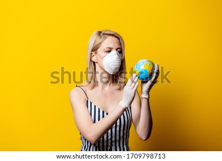 Blonde woman in face mask and gloves holds Earth globe on yellow background
