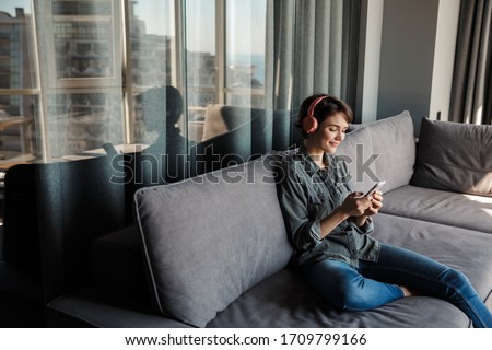 Image of nice young smiling woman using smartphone and headphones while sitting on couch at living room Royalty-Free Stock Photo #1709799166