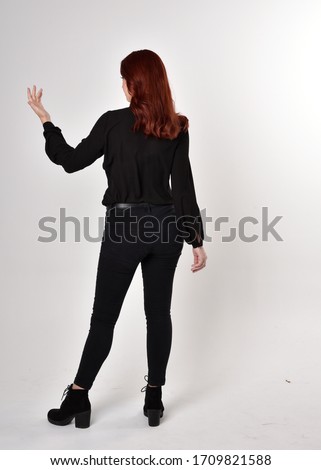 Portrait of a pretty girl with red hair wearing black jeans, boots and a blouse.  full length standing pose on a studio background, with back to the camera.
