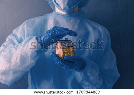 Doctor, a medical worker with medical gloves on his hands, a coat and a mask on his face, holds in his hands a small house with luminous windows.