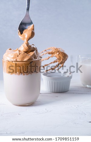 Spoon with whipped coffee falling into a Dalgona coffee on white background. Korean trendy drink. Vertical picture.