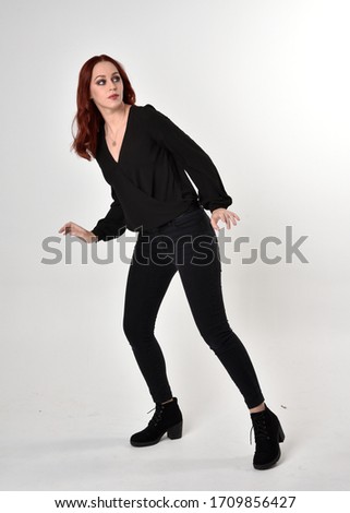 Portrait of a pretty girl with red hair wearing black jeans, boots and a blouse.  full length standing pose, facing the camera, on a studio background.