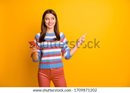 Portrait of positive cheerful girl promoter use smart phone point index finger copyspace indicate adverts promotion wear sweater trousers isolated over bright shine color background