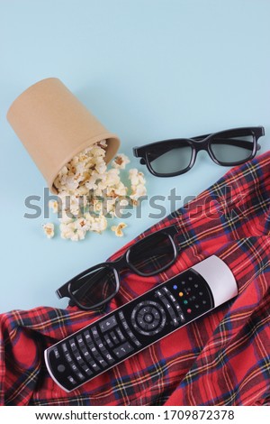 Watching at movie in quarantine concept.Home leisure composition. couple 3d glasses, craft papper cup of popcorn and TV remote control on a blue background and red plaid blanket.Top view, copy space Royalty-Free Stock Photo #1709872378