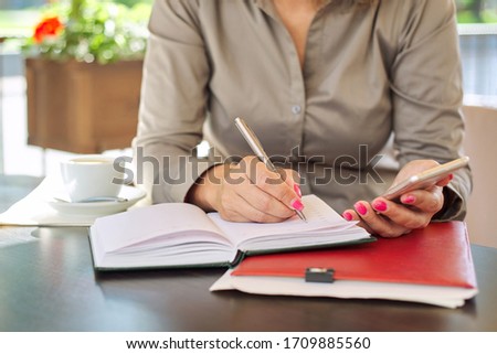 Businesswoman writing in business notebook using smartphone, close up, female sitting in summer outdoor cafe, coffee break