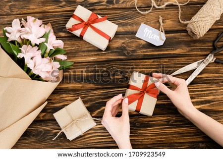 cropped view of woman touching ribbon on present near pink flowers wrapped in paper and tag with mom lettering, mothers day concept
