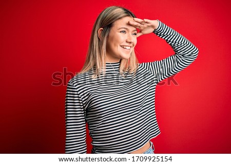 Young beautiful blonde woman wearing casual striped sweater over red isolated background very happy and smiling looking far away with hand over head. Searching concept.