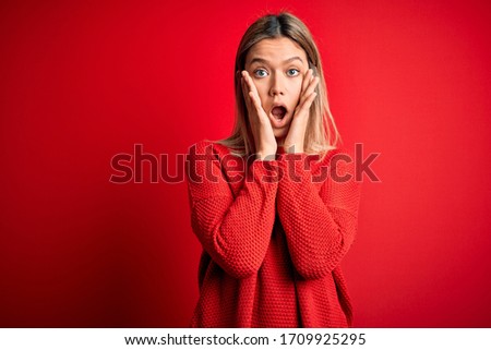 Young beautiful blonde woman wearing casual sweater over red isolated background afraid and shocked, surprise and amazed expression with hands on face