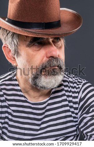Pensive, self-absorbed elderly overgrown man with a beard and mustache in stripped vest and cowboy hat