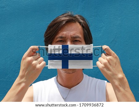 A man with Finland flag on hygienic mask in his hand and lifted up the front face on blue background. Tiny Particle or virus corona or Covid 19 protection. Concept of Combating illness.