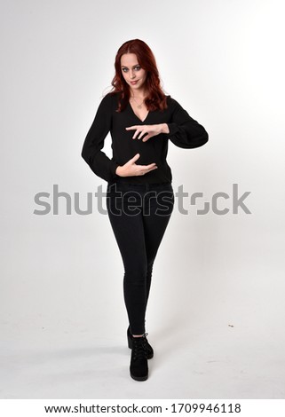 Portrait of a pretty girl with red hair wearing black jeans, boots and a blouse.  full length standing pose, facing the camera with a on a studio background.