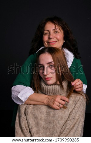 Mom and daughter . Beautiful woman 60 years old brunette and girl hugging. They are in sweaters on a black background in the studio. Emotional family photo