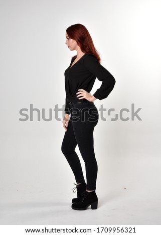 Portrait of a pretty girl with red hair wearing black jeans, boots and a blouse.  full length standing pose in side profile,  on a studio background.