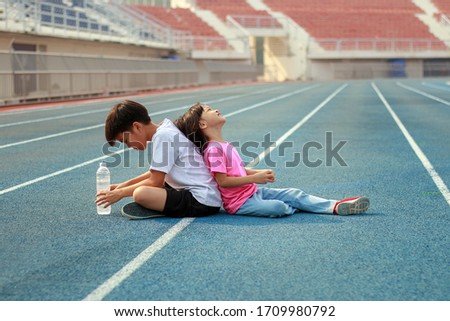 Boys and girls aged 4 to 6 years are sitting on the public open field for exercise. The girl was sitting next to the back of the boy after jogging on the blue treadmill. A bottle of water in the hand