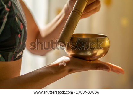 Young yogin woman holding a tibetan bowl and a stick against the beautiful and gentle light of morning during a yoga session, creating a meditative state through music. Theta binaural healing sounds. Royalty-Free Stock Photo #1709989036