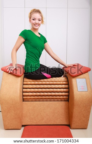 Smiling blond girl in sportwear sitting on modern relax massage equipment. Young woman doing relaxation exercise in healthy spa salon.