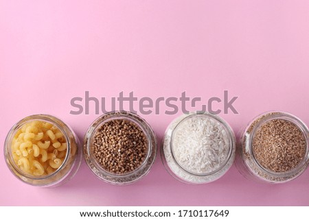 Storage of rice, buckwheat, wheat groats and pasta in glass jars. Crisis food stock for quarantine isolation period on pink background, Top view, Copy space