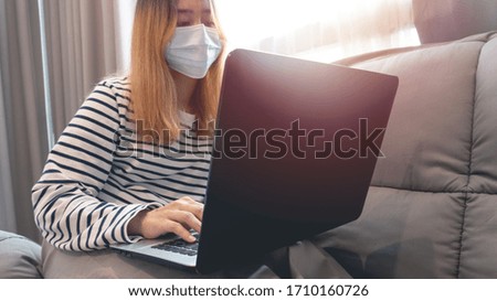 Women wearing protective blue face mask and working from home or learning online education on laptop computer while sitting on the grey sofa at the living room in during the Coronavirus or Covid-19.