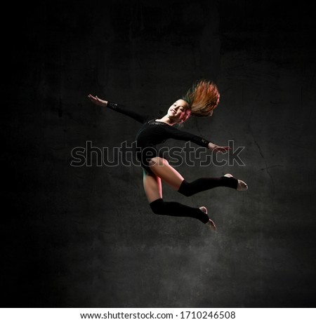 Cool sporty girl gymnast in black sport body and uppers jumping and making dymnastic pose in air over dark background. Rhytmhic gimnastics beauty cocnept