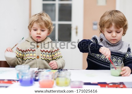 Two little twins boys having fun indoor, painting with different paints colors. Selective focus on one boy.