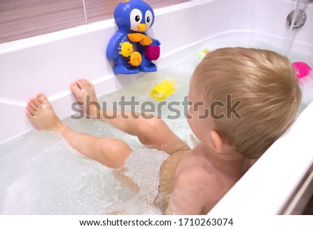 little boy with blond hair, 2 years old, sits in a bathtub filled with water and foam. Home furnishings, quarantine rest, relaxation, water treatments