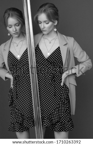 Young beautiful elegant woman in black and white polka dot dress and beige blazer standing and leaning on mirror. Fashion style shoot with reflection. Female model wearing jewelry earrings