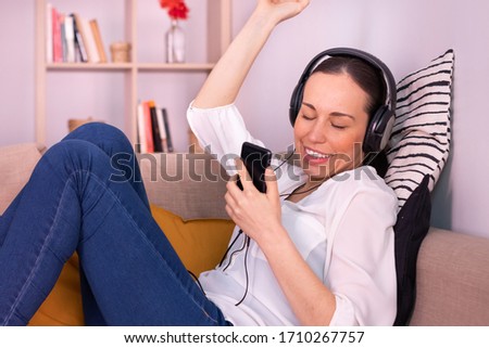 Happy pretty brunette young woman dancing with her smartphone stretched out on the sofa at home listening to music through headphones. She is wearing a white shirt and blue jeans.