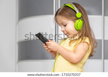 a little girl 3-4 years old stands in profile and listens to music in green headphones, in the hands of a smartphone. gadget addiction