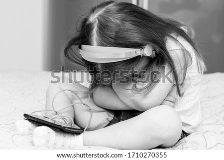 little girl sitting on the bed with big headphones with a smartphone. Offended pose, face hidden, monochrome. quarantine and self-isolation in connection with the caronavirus COVID-19