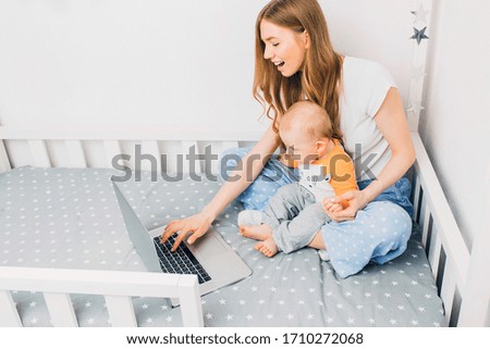 A young mother in pajamas with a small child, sitting on the bed, with a portable laptop. The concept of remote work in quarantine