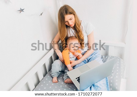 A young mother in pajamas with a small child, sitting on the bed, with a portable laptop. The concept of remote work in quarantine