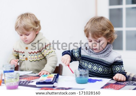 Two little twins boys having fun indoor, painting with different paints colors