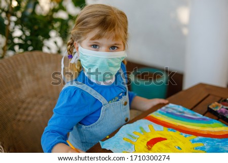 little toddler girl in medical mask painting rainbow with water colors during pandemic coronavirus quarantine disease. Children painting rainbows around the world with the words Let's all be well.