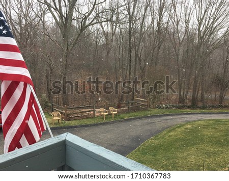 View off of a wooden deck at snow falling with the American flag and driveway in the background 
