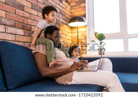 Big daddy with children sitting at home on the couch working remotely on a laptop