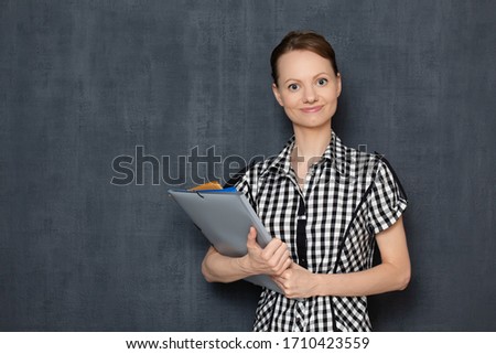 Studio portrait of happy friendly cheerful young blond woman wearing checkered shirt, smiling joyfully, holding colored folders, being ready to work, standing over gray background, copy space on left