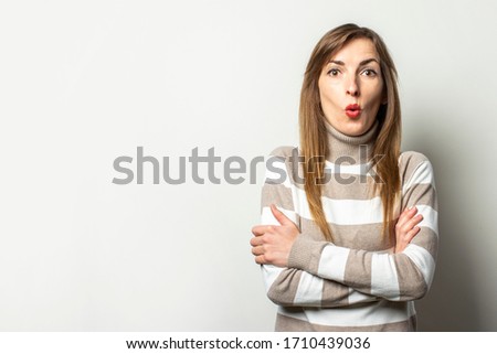 Portrait of a young friendly woman in a striped sweater hugged herself with a surprised face on an isolated light background. Emotional face. Gesture is cold, surprise, scary