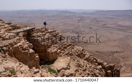 A male hiker standing on a lookout point at edge of Makhtesh Ramon Crater, in the Ramon Nature Reserve, city Mitzpe Ramon, Israel. Panoramic view on a dry wadi, sandy hills and desert rocky formations Royalty-Free Stock Photo #1710440047