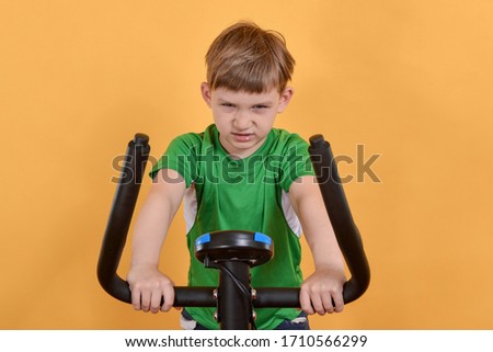 Portrait of a child on an exercise bike, the boy is engaged in cardio training and looking at the camera, on a yellow background.