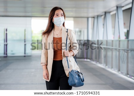 Young Asian professional business woman in casual shirt and medical mask during covid-19 or corona virus at modern walkway of Sky train station nearly office building. Bangkok, Thailand.