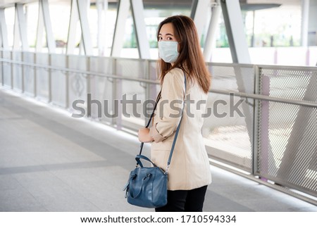 Young Asian professional business woman in casual shirt and medical mask during covid-19 or corona virus at modern walkway of Sky train station nearly office building. Bangkok, Thailand.