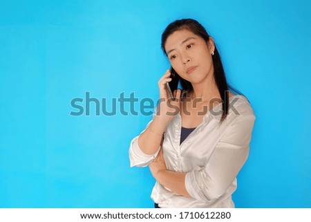 A serious looking Asian woman making a call on her cellphone, feeling upset and angry.  Plain light blue background. 