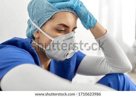 medicine, healthcare and pandemic concept - sad young female doctor or nurse wearing face protective mask or respirator for protection from virus disease holding to head