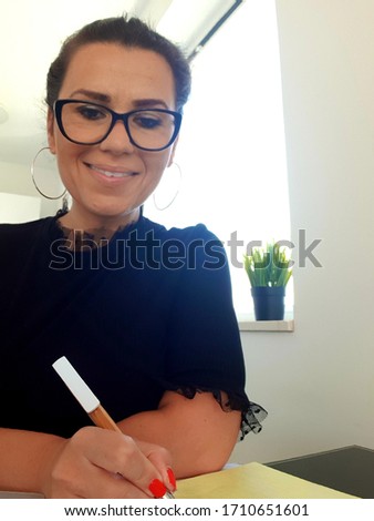 Cheerful smiling woman wearing eyeglasses  working at  computer and writing in a notebook sitting at the   kitchen at home . Working from home concept.