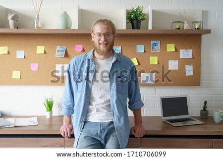 Smiling confident millennial business man remote worker look at camera stand at office desk work study from home. Happy male student professional designer freelancer having distance job portrait.