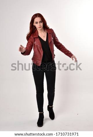  Portrait of a pretty girl with red hair wearing black jeans and boots with leather jacket.  full length standing pose with hand gesture on a studio background.