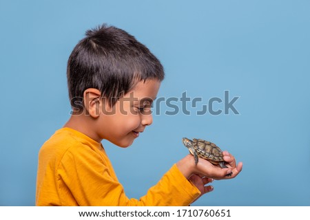 Shot of a young smiling boy wearing yellow shirt holding a turtle in his hand on a blue background with copy cpace.  The concept of children's hobbies. Happy childhood concept