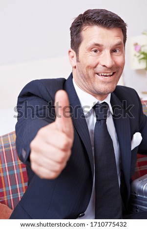 Laughing businessman holds thumbs up on sofa with suitcase